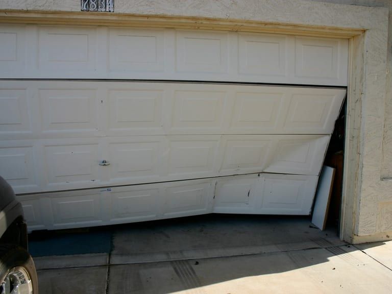A white garage door buckled off its track, panels bent and a gap open at one side.